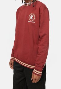 Persona che indossa una felpa bordeaux con polsini e orlo color crema, caratterizzata da un piccolo logo Karl Kani sul petto, abbinata a pantaloni neri.