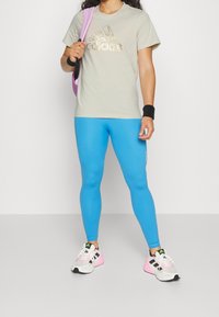 Lys grå t-shirt med guld Adidas-logo, blå leggings og hvide sneakers med pink accenter. Accessories inkluderer en lilla rygsæk og håndledsarmbånd.