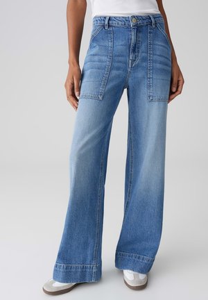 Jean en denim bleu à jambes larges avec de grandes poches plaquées devant et des ourlets retroussés, porté avec des baskets blanches et un haut blanc.