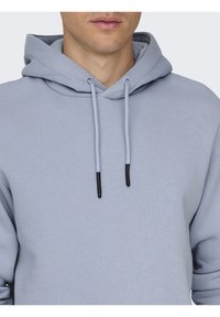 Sudadera azul claro fabricada en tejido suave, con un bolsillo frontal, cordones ajustables con puntas negras y un corte relajado.