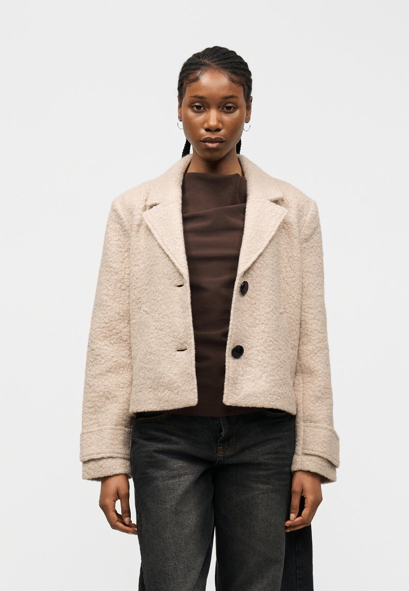 Γυναίκα με πλεξούδες που φοράει μπεζ υφή cropped παλτό, σκούρο καφέ τοπ και σκούρα ξεβαμμένα τζιν μπροστά σε απλό φόντο.