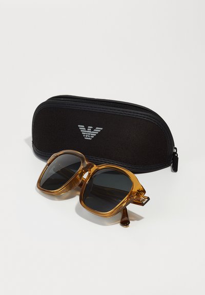 Lunettes de soleil carrées avec des montures teintées ambre et des verres foncés, à côté d'un étui rigide noir arborant un logo en argent. Surface de l'étui texturée.