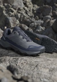 Σκούρα γκρι και μαύρα πεζοπορικά παπούτσια Adidas Terrex σε βραχώδες έδαφος, που δείχνουν πλευρά και σόλα με ανθεκτικό πέλμα.