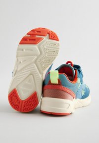 Buty sportowe z niebieską cholewką i czerwonymi akcentami oraz gumową podeszwą. Podeszwa zewnętrzna ma teksturowany wzór i zawiera jasnopomarańczowy wkład.