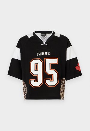 Férfi fekete, túlságosan bő sportmezben, rajta az 95-ös szám, és erősen szakadt térd alatti rövidnadrágban, kezét zsebében tartva áll.