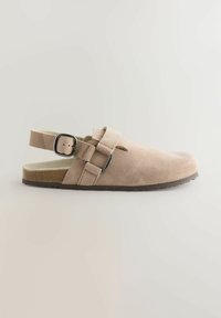 Chaussure beige en daim à enfiler avec une sangle à boucle ajustable, un bout rond et une semelle en caoutchouc texturé. Design minimaliste avec des découpes.