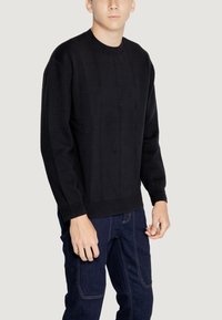 Maglione nero a maglia con un motivo a griglia testurizzato, collo rotondo e polsini a coste. Indossato sopra jeans scuri, presenta un discreto dettaglio del logo.
