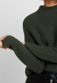 Person trägt einen dunkelgrünen, gerippten Strickpullover mit langen Ärmeln, ein Arm ausgestreckt, und schwarze Hosen mit sichtbarem Reißverschluss und Knöpfen.