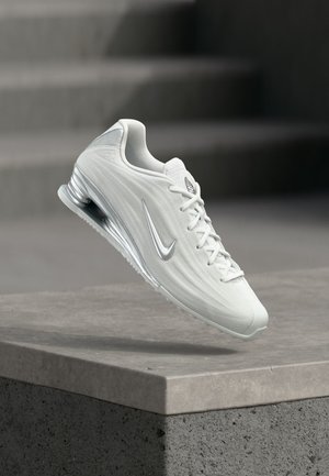 Hvid Nike-sneaker med sølv swoosh-logo, luftpolstret sål og snørebånd, vist svævende over en betonblok.