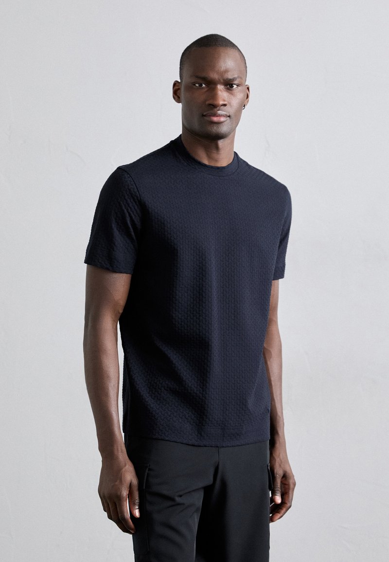 Σκούρο μπλε κοντομάνικο t-shirt με υφή και λαιμόκοψη crew neck, που διαθέτει γεωμετρικό σχέδιο και ευθύ στρίφωμα. Φορεμένο με μαύρο παντελόνι.