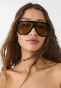 Lunettes de soleil noires surdimensionnées avec une monture géométrique et des verres teintés marron. Le style est audacieux avec une finition en plastique lisse.