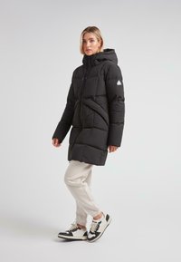 Kvinne iført en svart quilted hettejakke, lysebeige joggebukser og hvite og svarte sneakers som står sidelengs foran en ensfarget bakgrunn.