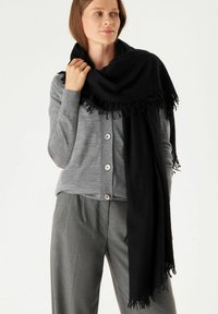 Écharpe noire avec des accents de frange drapée autour d'un modèle, associée à un cardigan gris et à un pantalon gris ajusté. Matériau léger et doux.