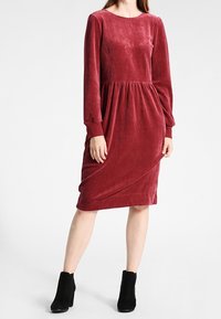 Czerwona sukienka z weluru z długimi rękawami, okrągłym dekoltem i marszczonym stanikiem. Sylwetka w kształcie litery A, długość do połowy łydki, zestawiona z czarnymi botkami za kostkę.