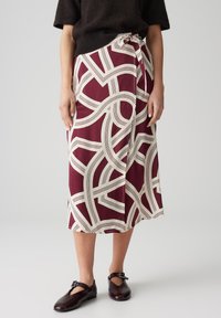 Jupe midi bordeaux avec un motif crème abstrait, dotée d'un nœud sur le côté et en tissu doux. Associée à des chaussures plates marron foncé.