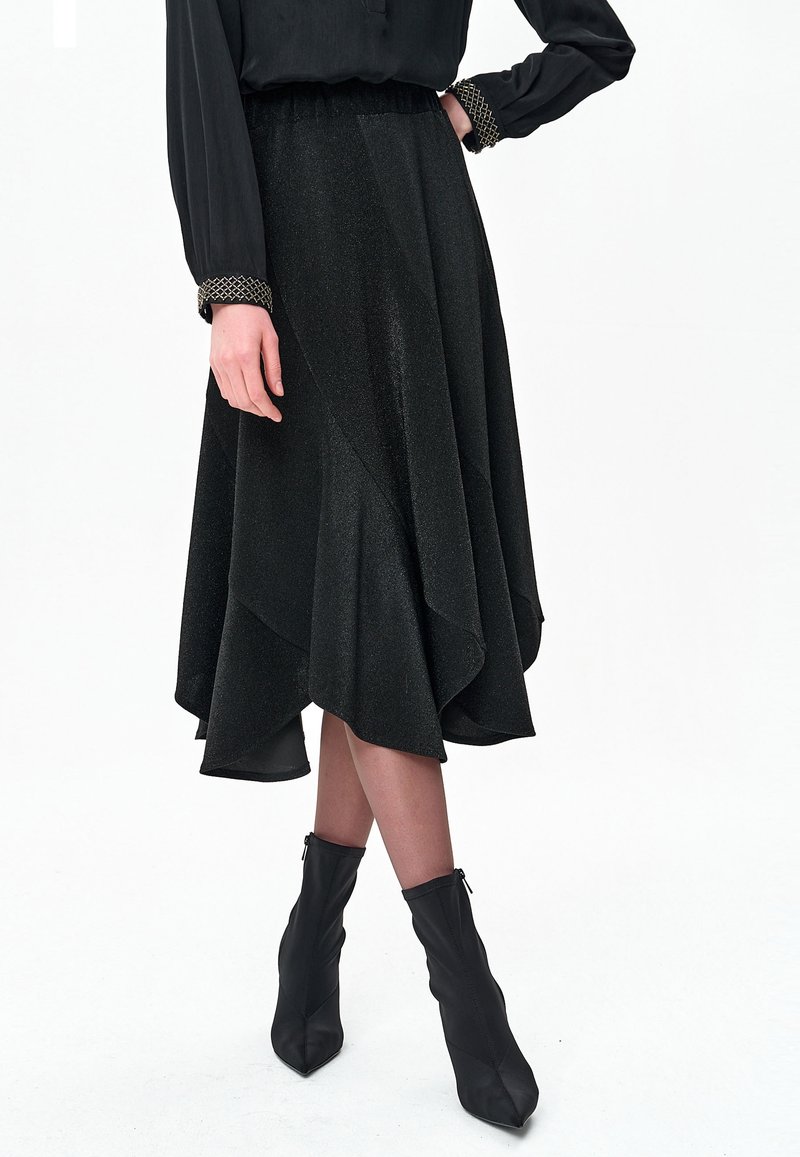 Czarna asymetryczna spódnica o teksturowanym, błyszczącym wykończeniu i warstwowym kroju, z marszczonym paskiem w talii. Noszona z dopasowanymi czarnymi botkami za kostkę.