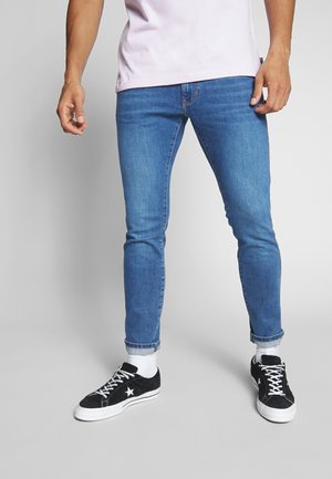 Jeans de tiro bajo color azul claro con un acabado desgastado, arremangados en los tobillos. Llevados con zapatillas de ante negras y calcetines blancos.