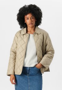 Veste matelassée beige avec une coupe légèrement oversized, boutons-pression et col classique, portée sur un léger pull et une jupe en denim.