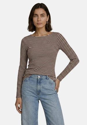 Femme aux cheveux foncés portant des boucles d'oreilles créoles dorées, un t-shirt à manches longues rayé marron et blanc, et un jean bleu clair, une main sur la hanche.