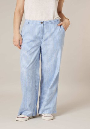 Femme portant un pantalon large à rayures verticales bleu clair et blanc et des baskets blanches avec un haut blanc, debout une main dans la poche.