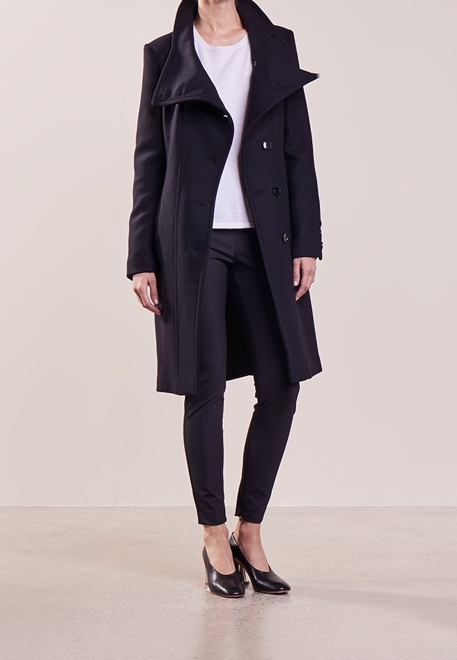 Femme portant un manteau noir à double boutonnage, un haut blanc, un pantalon noir ajusté et des talons noirs, debout sur un fond neutre.