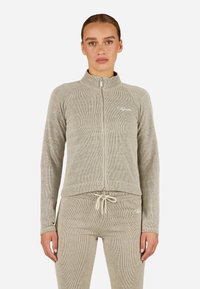 Gai beige krāsas jaku ar rāvējslēdzēju, ar ribotu tekstūru, augstu apkakli un logo akcentu; savienota ar atbilstošām pieguļošām biksēm.