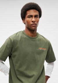 T-shirt de algodão verde-oliva com decote redondo e mangas curtas, com o texto "EARLY BIRDS" em laranja no lado esquerdo.