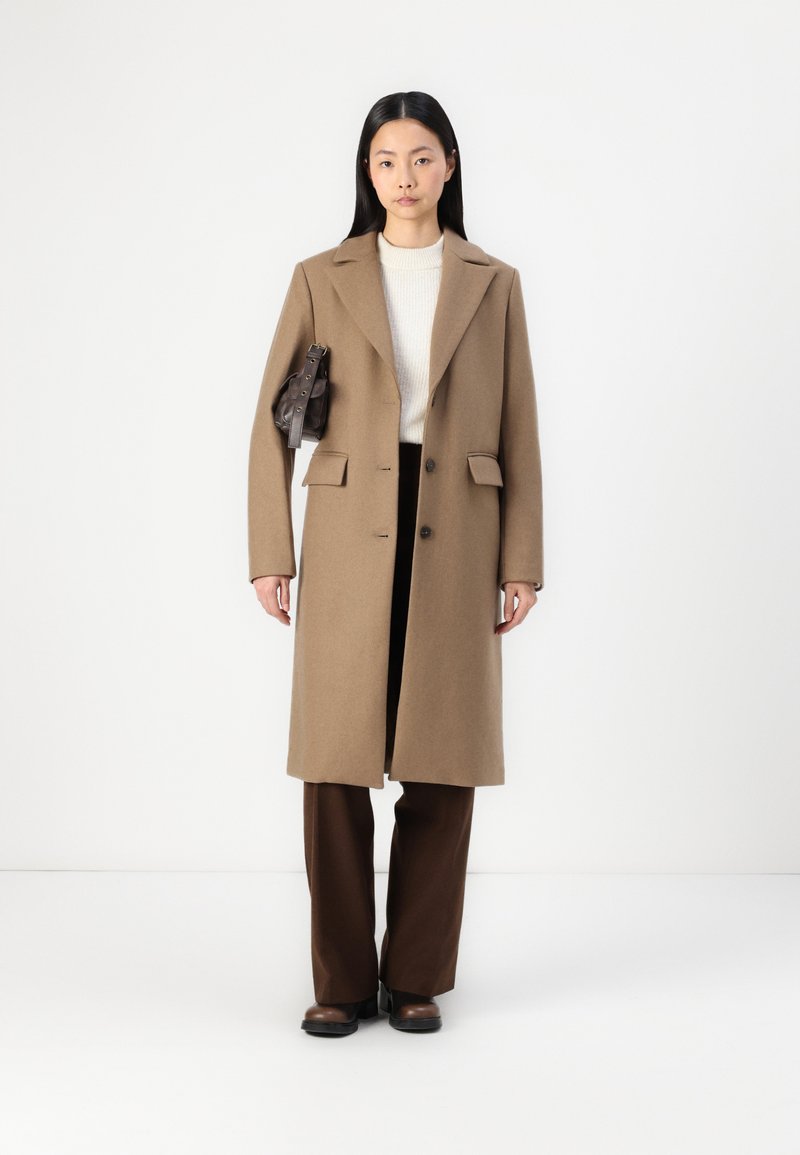 Lungo cappotto in lana color cammello con colletto a revers, due tasche frontali e due bottoni, abbinato a pantaloni marrone scuro e una piccola borsa in pelle.