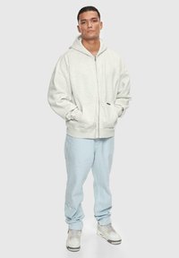 Lys grå zip-up hættetrøje med stor hætte, forlommer og løs pasform, parret med lyseblå jeans og hvide sneakers.