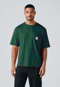 Mand iført en mørkegrøn kortærmet T-shirt med en lille brystlomme og sorte cargo-bukser foran en lys grå baggrund.