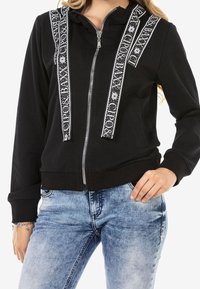 Black zip-up hoodie with white patterned accents along the front, featuring a drawstring hood and ribbed cuffs. Worn with light blue jeans.