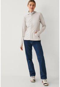 Mujer de pie con una chaqueta con capucha de color gris claro con cremallera, jeans azules y zapatillas blancas sobre un fondo claro y liso.