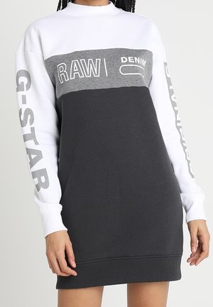 Kobieta nosząca sukienkę z długim rękawem w kolorowe bloki z napisem "RAW DENIM" na piersi i tekstem "G-STAR" na obu białych rękawach.