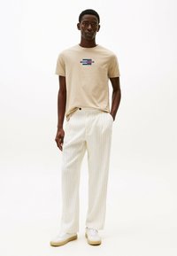 Camiseta de algodón beige con logo gráfico en el pecho, combinada con pantalones de pierna ancha blancos con rayas. La modelo lleva zapatillas blancas.