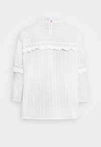 Vit långärmad blus med texturerat prickmönster, spetsdetaljer, volang på axlarna och knäppt nyckelhål vid den höga kragen, sedd bakifrån.