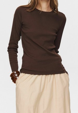 Kvinde iført en tætsiddende brun langærmet top, beige nederdel med lommer og stablede brune armbånd på venstre håndled mod hvid baggrund.