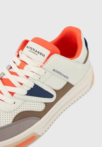 Sneaker bianca con un mix di texture, tra cui scamosciato e rete, con dettagli arancione, inserti blu navy e una suola in gomma spessa.