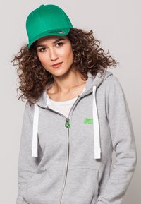 Gorra verde con visera curva, llevada con una sudadera gris con cremallera que cuenta con cordones blancos y un pequeño logo verde en el pecho.