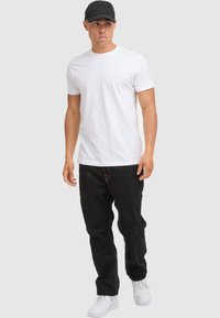 Un bărbat îmbrăcat cu o sapcă neagră, un tricou alb, pantaloni negri cu cusături albe și adidași albi, stă în picioare pe un fundal deschis, simplu.