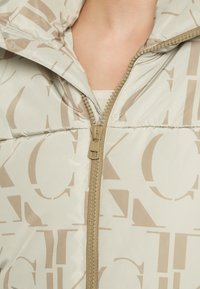 Vadderad jacka i ljusbeige med en subtil glänsande finish, med ett brunt logomönster, hög krage och en frontdragkedja med guldfärgade detaljer.