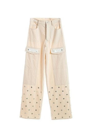 Joven mujer con pantalones y chaqueta cargo color crema con puntos plateados, camiseta gráfica beige y zapatos beige texturizados, de pie sobre fondo blanco.