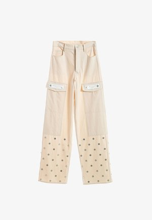 Joven mujer con pantalones y chaqueta cargo color crema con puntos plateados, camiseta gráfica beige y zapatos beige texturizados, de pie sobre fondo blanco.