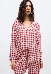 Γυναίκα που φοράει κόκκινη και λευκή πιτζάμα με μικρά σχέδια καρδιών, στέκεται μπροστά σε απλό λευκό φόντο.