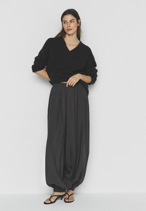 Sieviete stāv, rokas uz gurniem, vilkusi melnu V-veida izgriezuma džemperi, brīvas melnas bikses un melnas sandales ar papēdi, uz vienkārša fona.