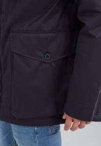 Svart jacka med stor knäppt ficklock på sidan, bärs över blå jeans, med synlig hand nära fickan.