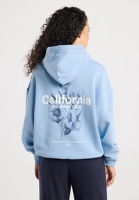 Frau mit lockigem Haar, die einen hellblauen Hoodie trägt, auf dem "California Botanical Club" und florale Grafiken auf dem Rücken zu sehen sind, kombiniert mit dunklen Hosen.