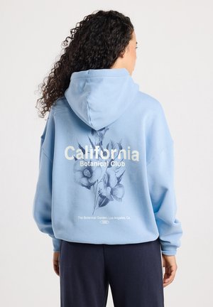 Mujer con cabello rizado que lleva puesta una sudadera azul claro con "California Botanical Club" y gráficos florales en la parte de atrás, combinada con pantalones oscuros.