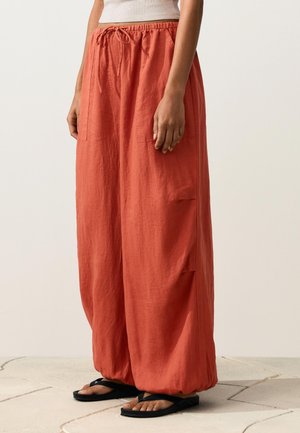 Persona che indossa pantaloni larghi color ruggine con coulisse e polsini raccolti, e sandali neri, in piedi su un pavimento piastrellato chiaro.