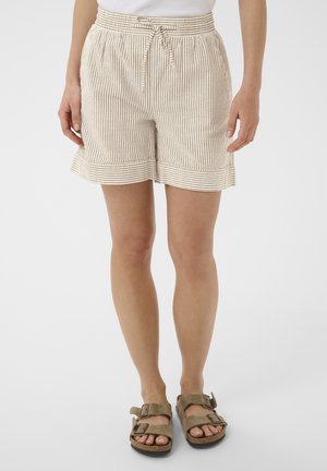 Donna che indossa pantaloncini beige e bianchi a righe con coulisse e sandali in camoscio marrone con fibbie, in piedi davanti a uno sfondo semplice.