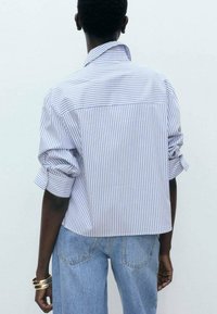 Chemise à rayures blanches et bleues avec boutons et manches retroussées, coupe droite. Portée avec un jean bleu clair à jambes larges.